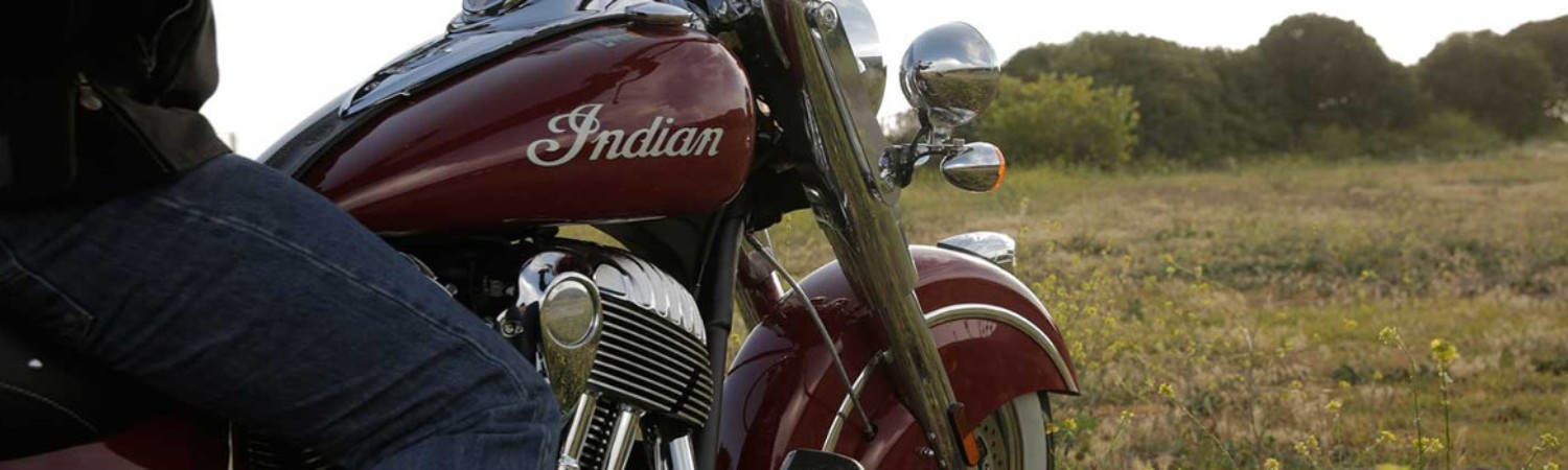 Close-up of person wearing jeans on a dark red Indian Motorcycle® overlooking an empty grassy field.